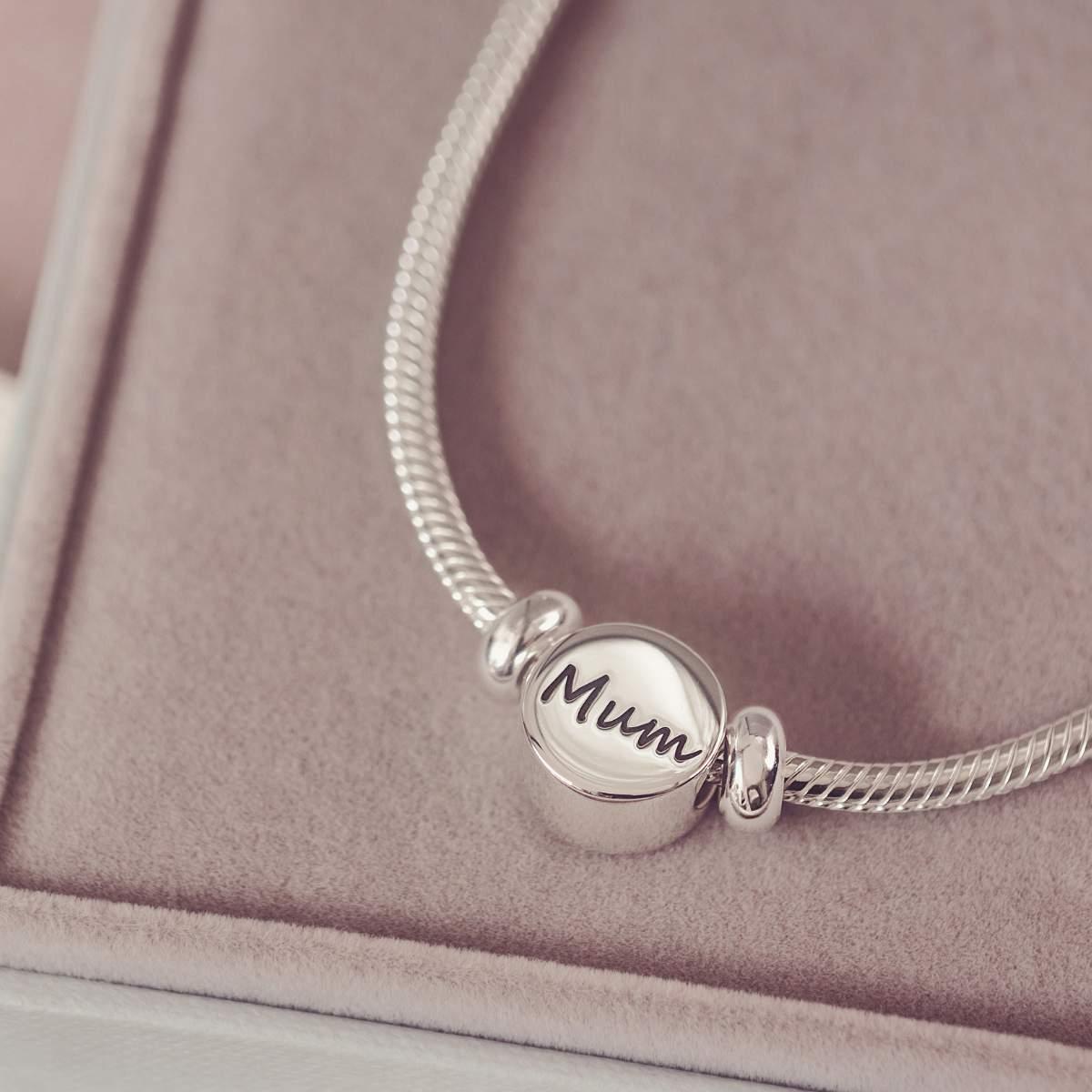 Silver bracelet with a round charm inscribed with 'Mum' on a pink surface