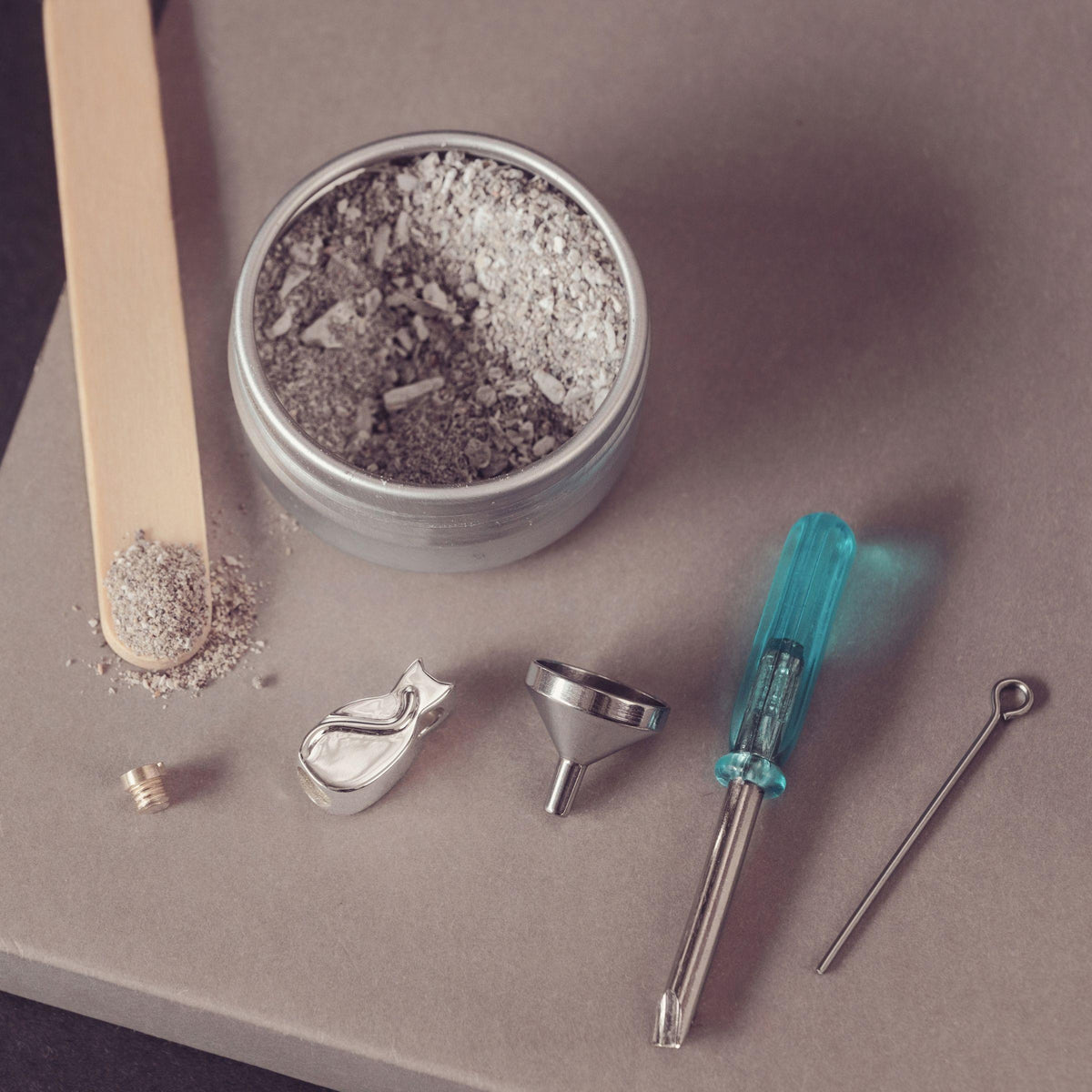 A metal tin filled with ashes, arranged on a taupe surface with a wooden spatula holding some ash, a small metal screw, a silver funnel, ametal piece, and a turquoise-handled tool.