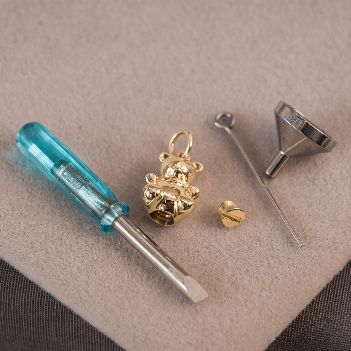 Gold bear-shaped charm and matching jewelry-making tools arranged on a beige textured surface, including a small screwdriver with a translucent blue handle, a metal funnel, and a pin.