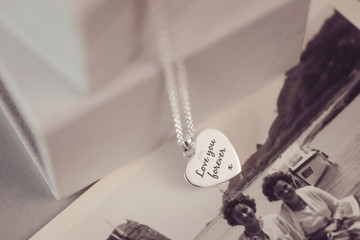 A silver heart-shaped pendant necklace engraved with 'Love you forever' resting on top of a sepia-toned photograph, with a white gift box blurred in the background.