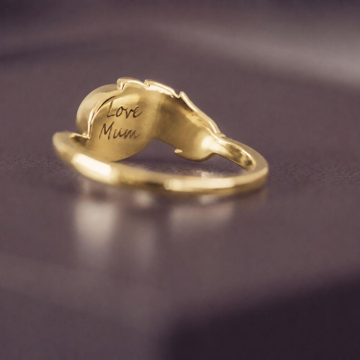 A gold ring shaped like a mother's hand holding a heart, with 'Love Mum' engraved on the heart, resting on a dark purple surface.