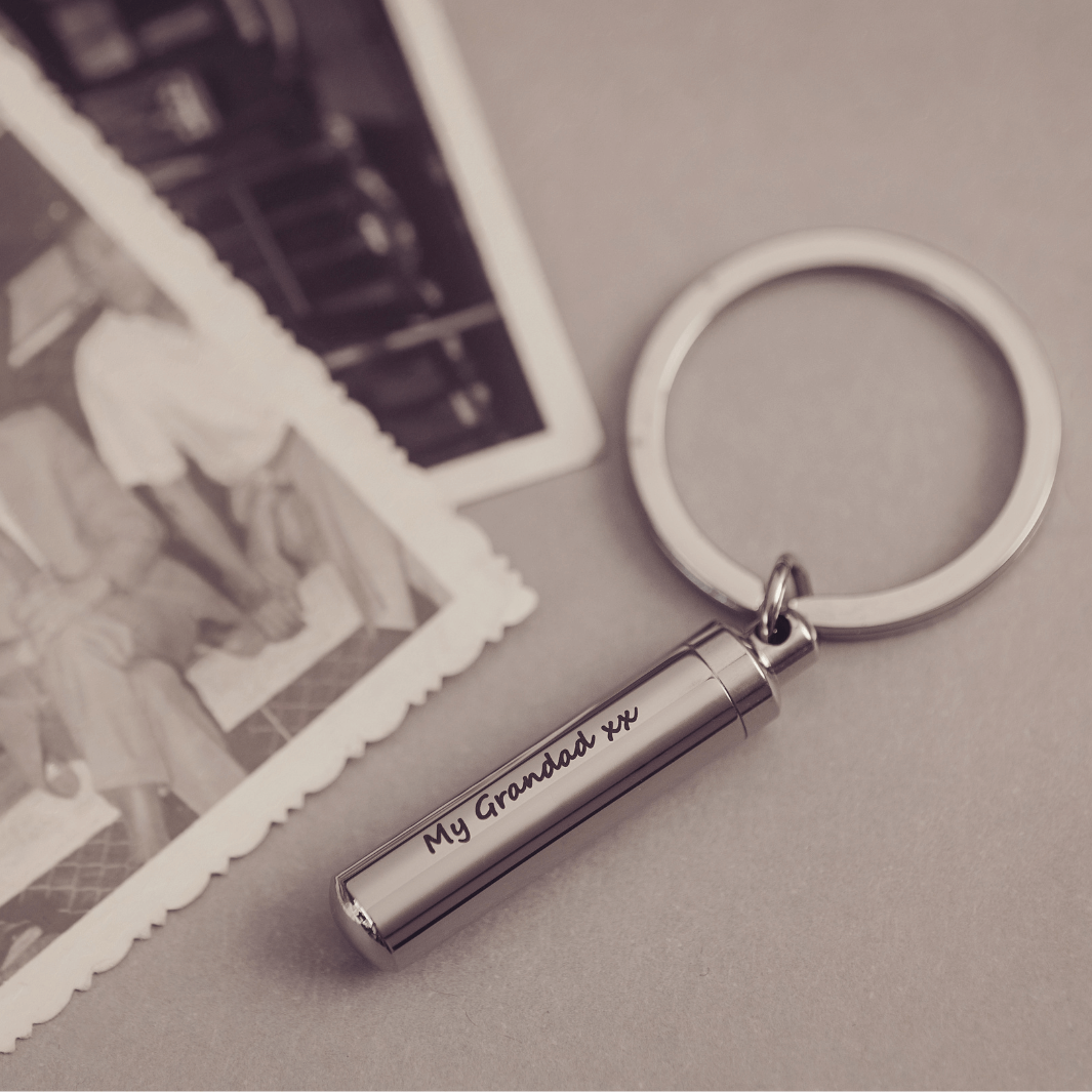 Cylindrical keyring which unscrews to hold cremation ashes or hair, engraved with the message "My Grandad xx" on one line, on a light grey background with vintage photos in the background.