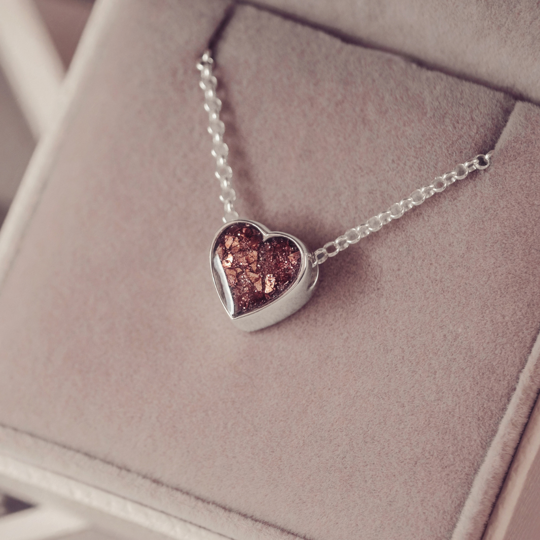 A silver heart pendant necklace with a textured, glittery brown center, resting on a dusty pink velvet jewelry box.