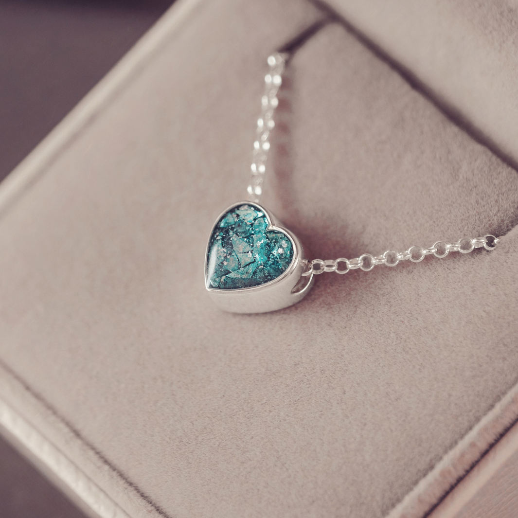 A silver heart-shaped pendant necklace with a textured blue and green mosaic glass center, resting on a beige jewelry box.