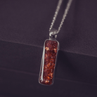 A sophisticated silver necklace featuring a rectangular pendant filled with shimmering orange crystal and resin, delicately layered with cremation ashes, showcased against a dark background, on a silver belcher chain.
