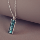 A sophisticated silver necklace featuring a rectangular pendant filled with shimmering turquoise crystal and resin, delicately layered with cremation ashes, showcased against a dark background, on a silver belcher chain.