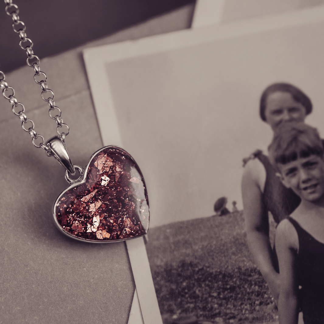 Sterling silver heart necklace with a central section filled with memorial ashes and brown crushed crystals, displayed on a beige fabric background next to a black and white photograph