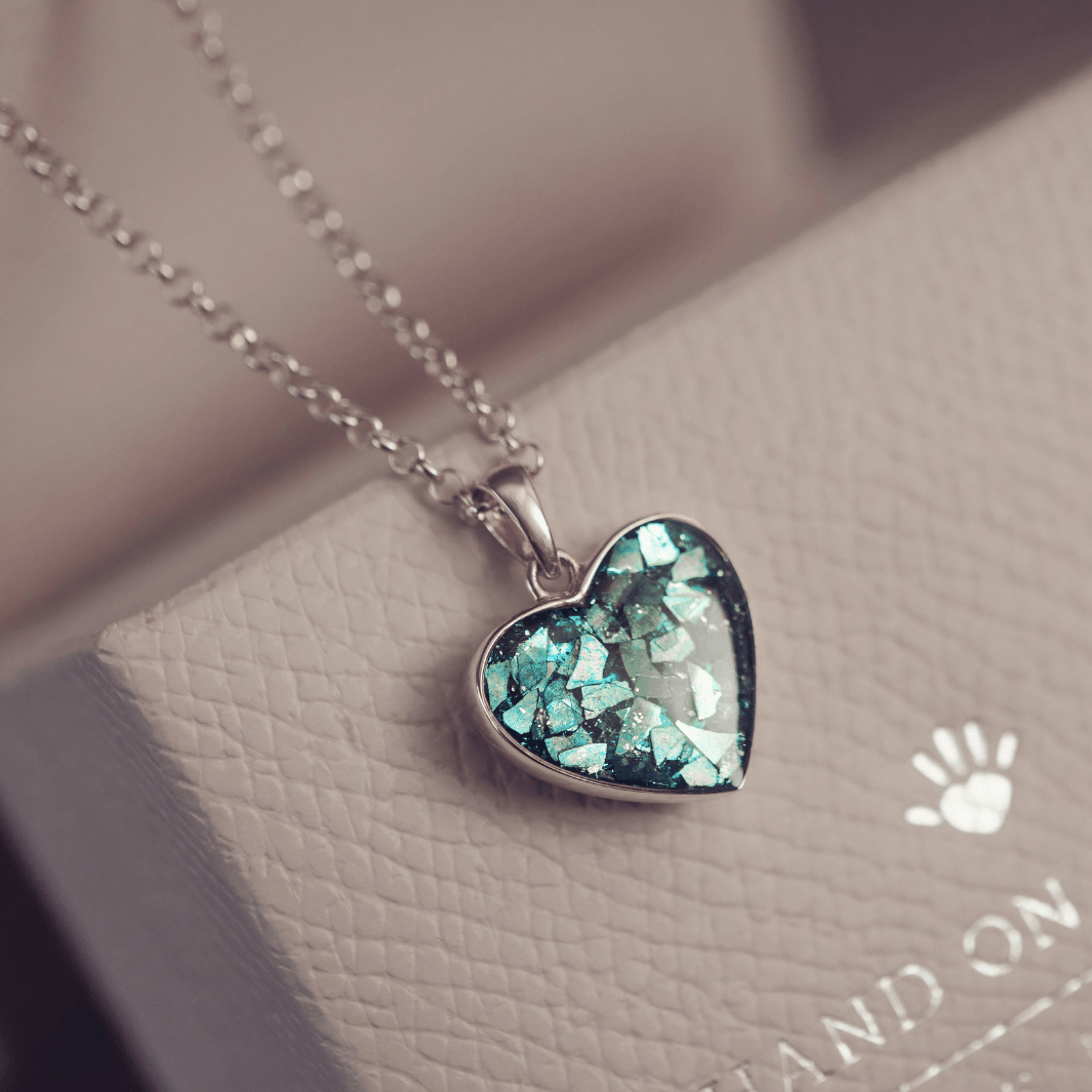 Sterling silver heart necklace with a central section filled with memorial ashes and turquoise crushed crystals, displayed on a grey textured jewellery box