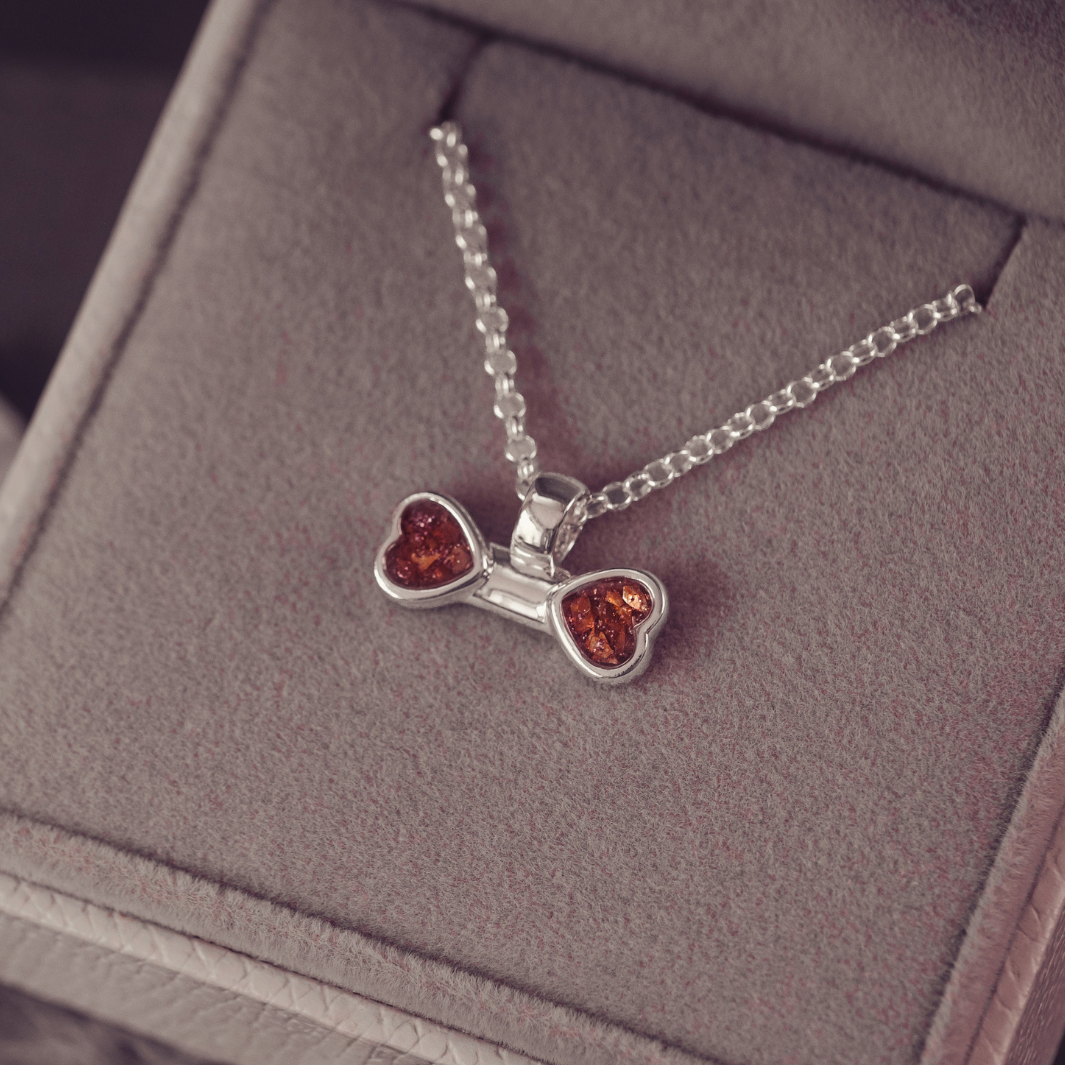 A silver necklace with a bow-shaped pendant featuring two amber heart inlays, displayed inside a gray jewelry box.