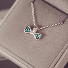 A silver necklace with a bone-shaped pendant featuring two heart-shaped blue-green crystal inlays, resting on a beige jewelry box.