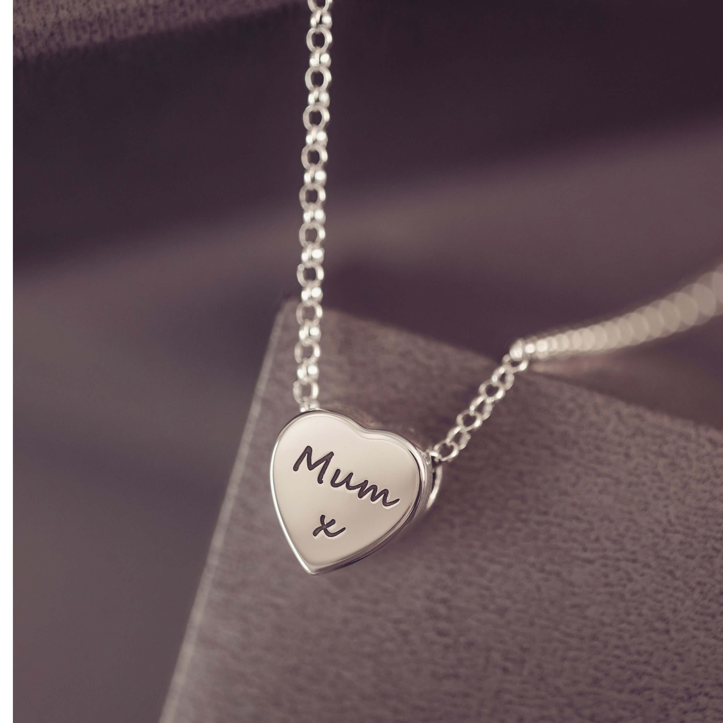 Silver heart-shaped pendant necklace with 'Mum x' engraving on a dark surface