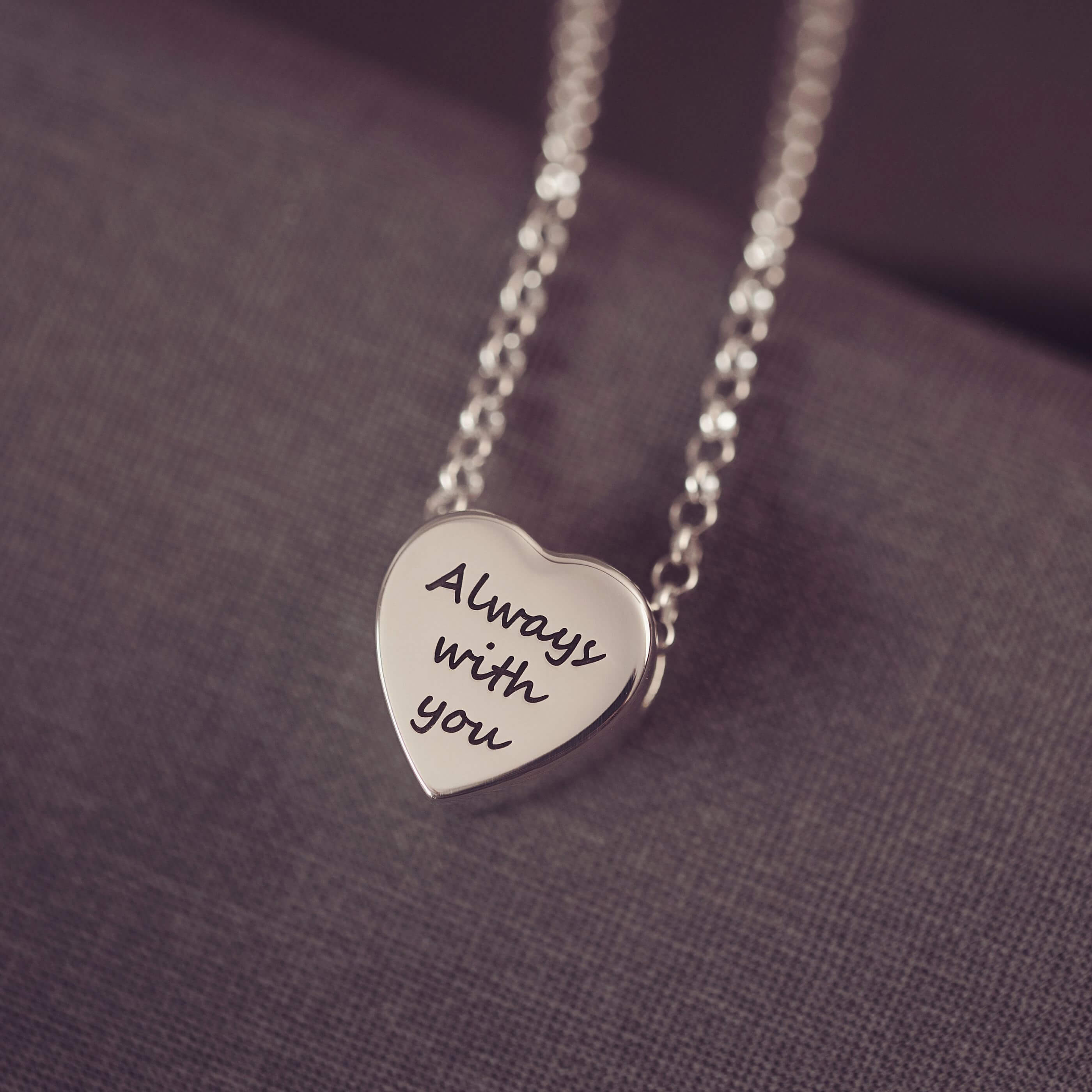 Silver heart-shaped pendant necklace with 'Always with you' engraving on a purple fabric background
