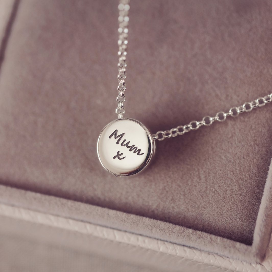 A silver round pendant necklace engraved with 'Mum x' in black script, resting on a dusty mauve velvet jewelry box.