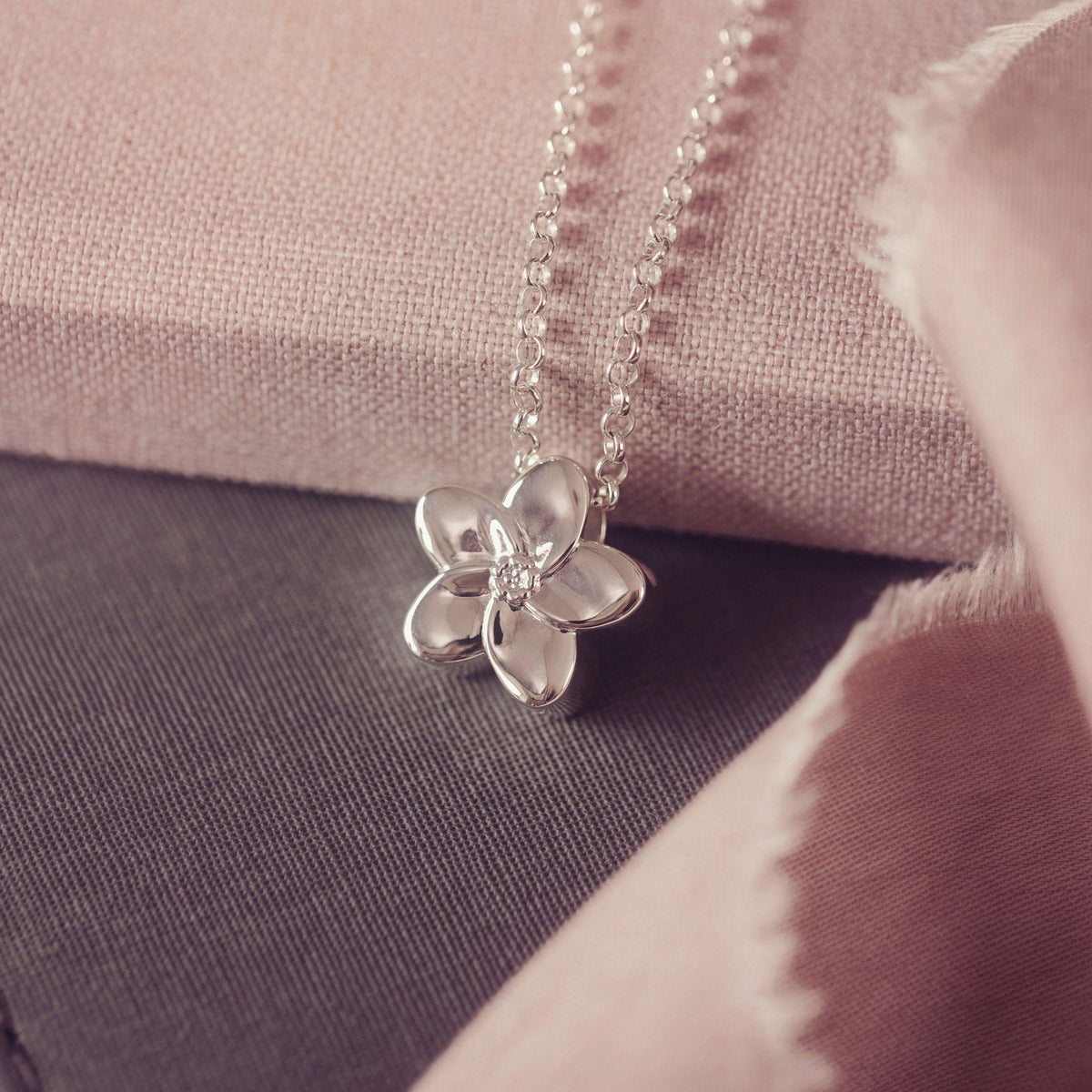 A silver flower pendant necklace resting on a textured pink fabric surface with a dark gray fabric background.