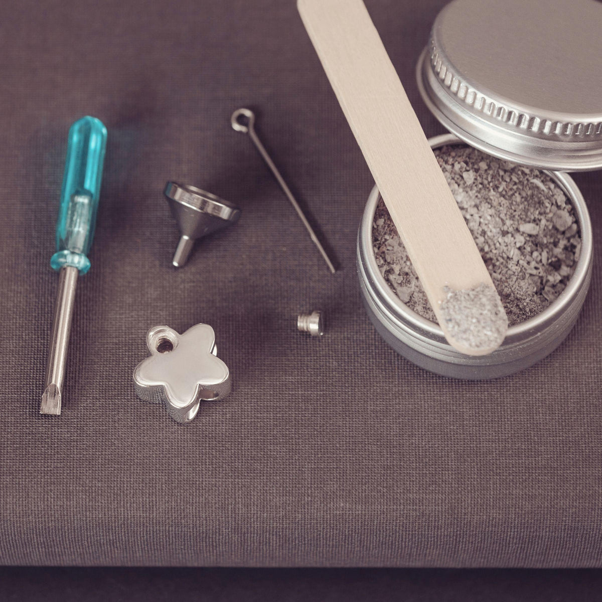Silver jewelry-making supplies arranged on a dark textured surface, including a small turquoise-handled screwdriver, metal funnel, pin, flower-shaped charm and an open tin of ashes with a wooden spatula.