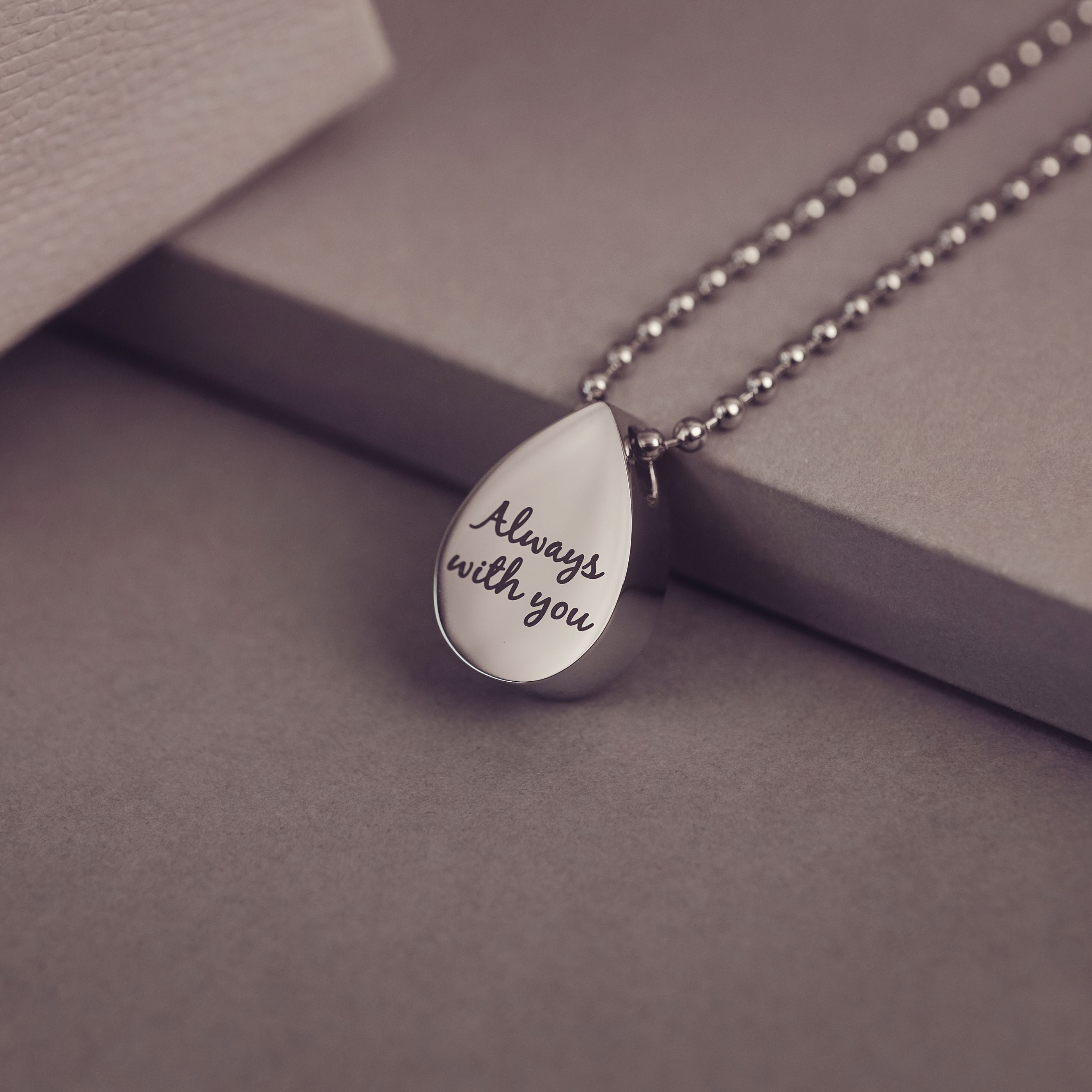 Silver teardrop pendant necklace with 'Always with you' engraving on a gray surface