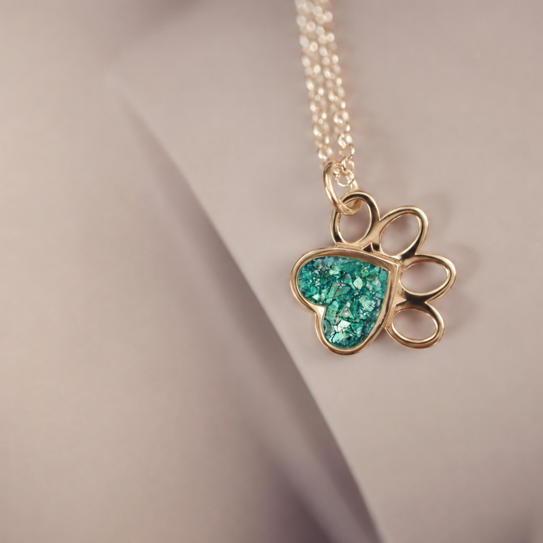 A gold paw print pendant necklace with a glittery turquoise heart inlay, resting on a neutral beige surface.