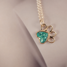 A gold paw print pendant necklace with a glittery turquoise heart inlay, resting on a neutral beige surface.