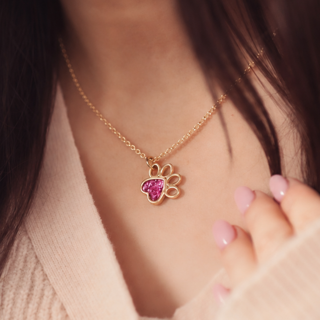 A close-up lifestyle shot of a gold chain necklace with a paw print pendant featuring a pink heart-shaped center, worn by a person in a beige knit top.