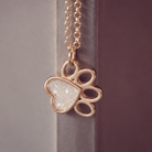 A rose gold paw print pendant necklace with a heart-shaped clear stone inlay, hanging against a dark gray and purple background.
