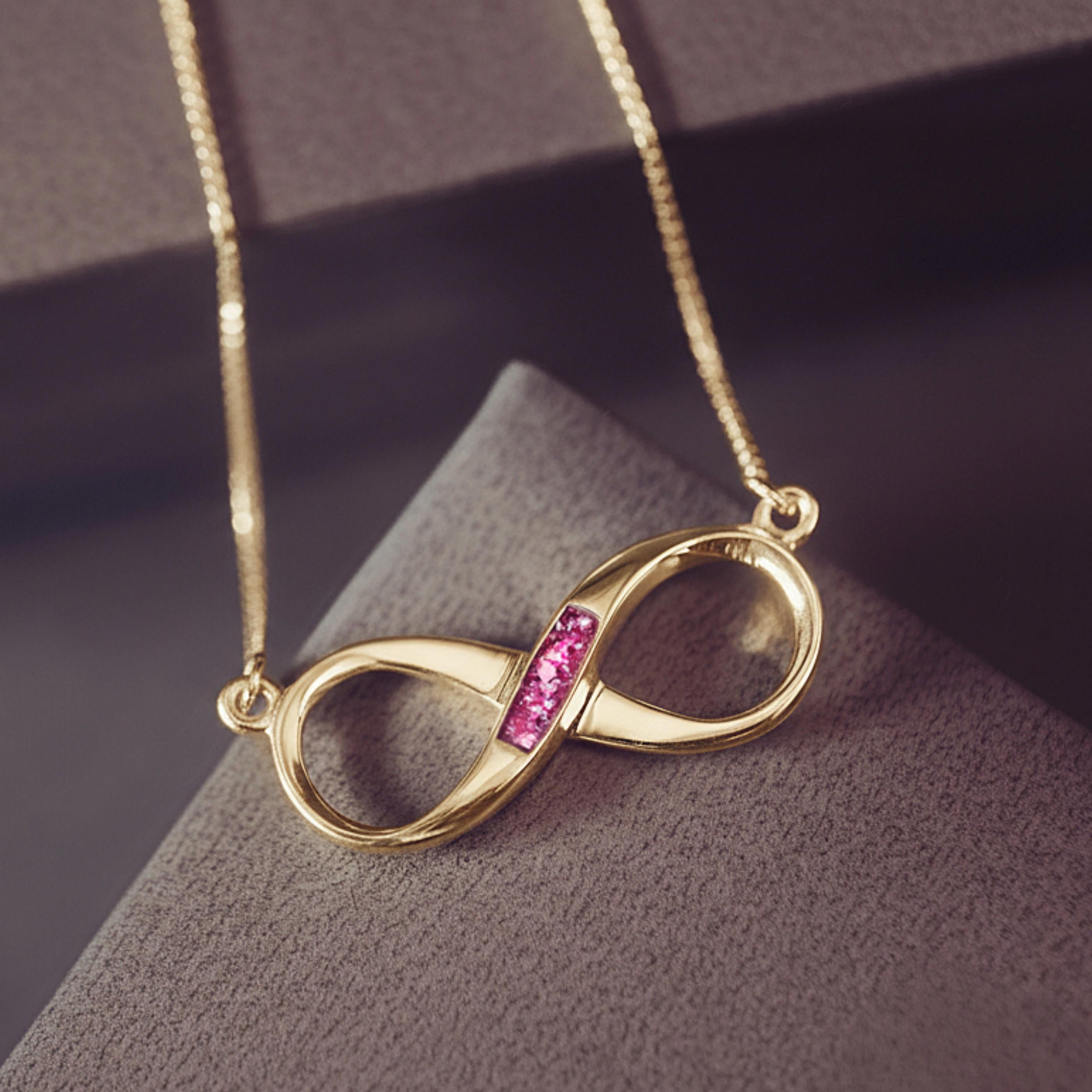 A gold Infinity shaped necklace with pink gemstones on a grey and purple box