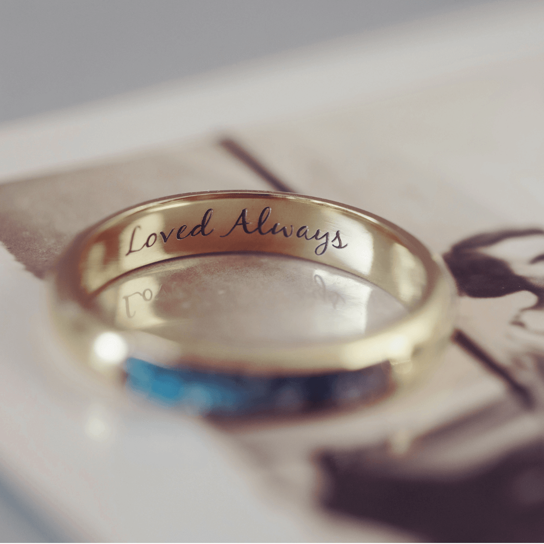 A gold ring with 'loved always' engraved. The ring is sitting on a black and white blurred photograph. 