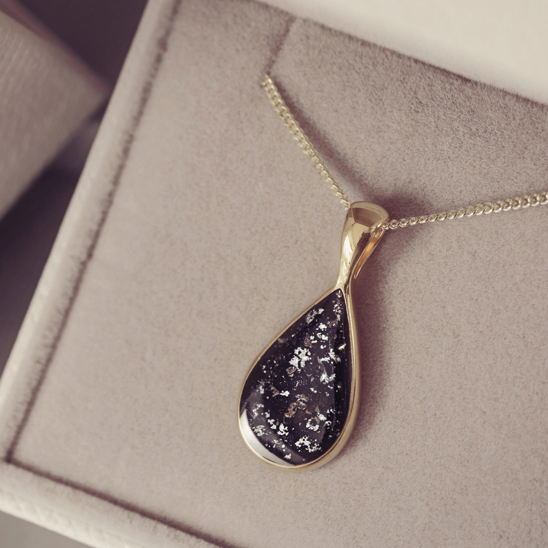 Gold chain with a teardrop-shaped pendant, including ashes in a black setting, on a soft surface
