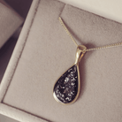 Gold chain with a teardrop-shaped pendant, including ashes in a black setting, on a soft surface