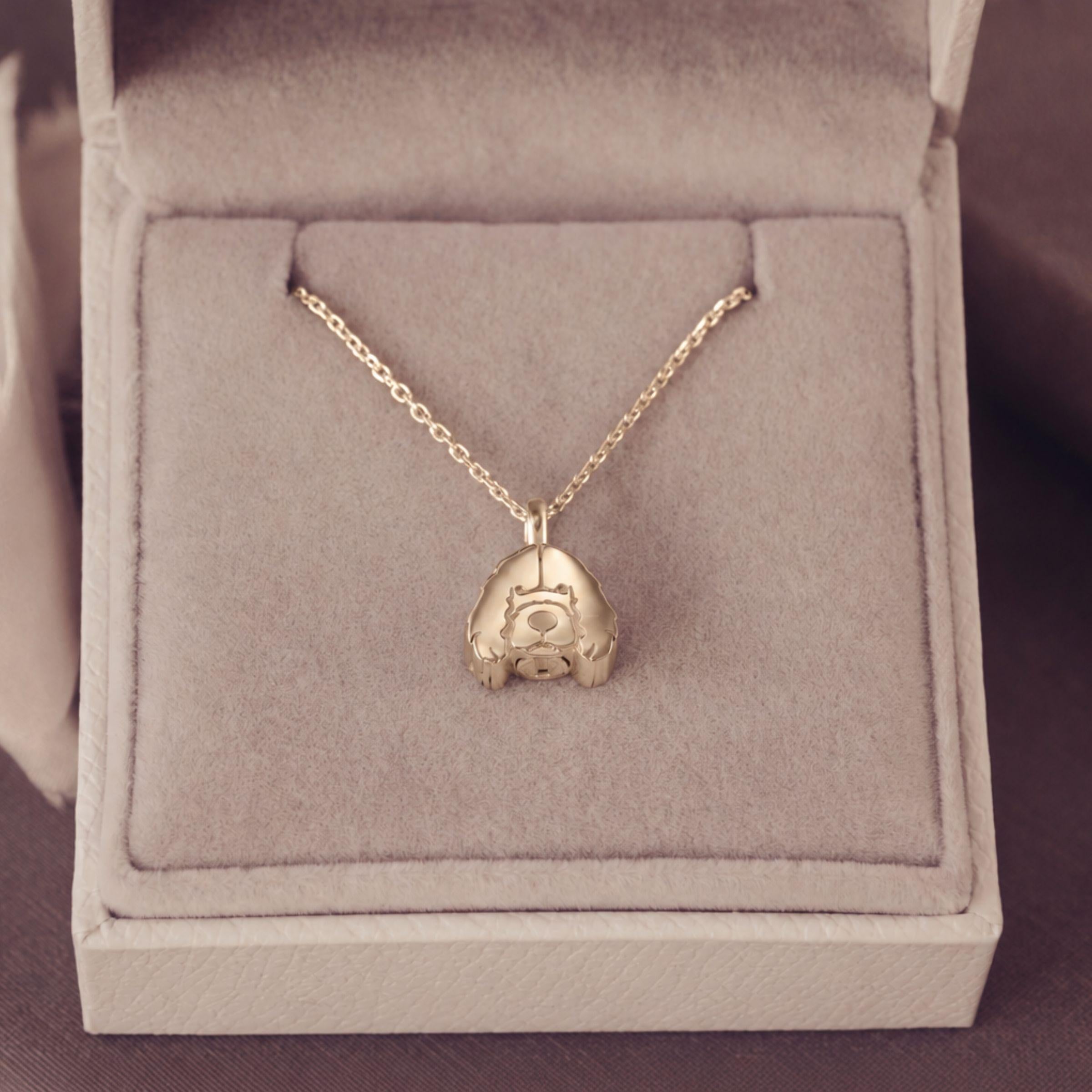 A gold pendant necklace displayed inside an open light beige jewelry box. The pendant is of a stylized dog, suspended from a delicate chain.