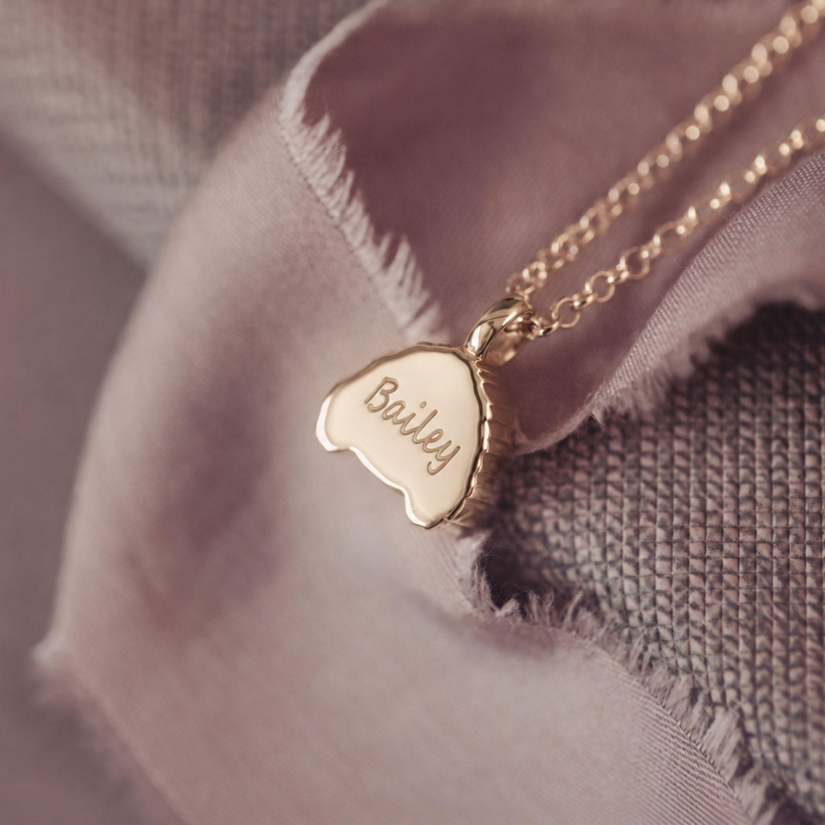 A close-up of a gold necklace with a small engraved pendant that reads 'Bailey', resting on soft dusty pink fabric and textured gray woven material.