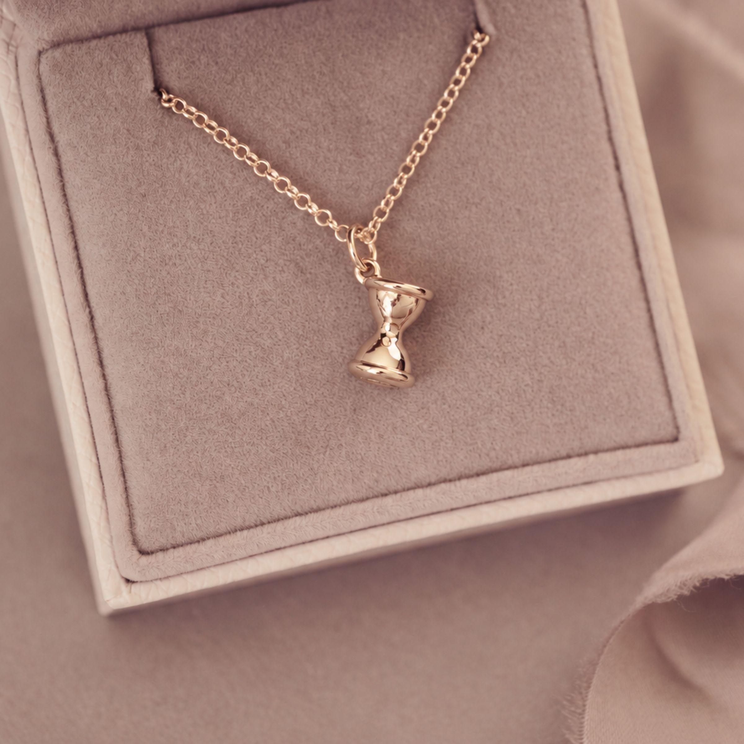 A gold chain necklace with a small hourglass pendant, displayed inside a beige jewelry box with a soft beige fabric background.