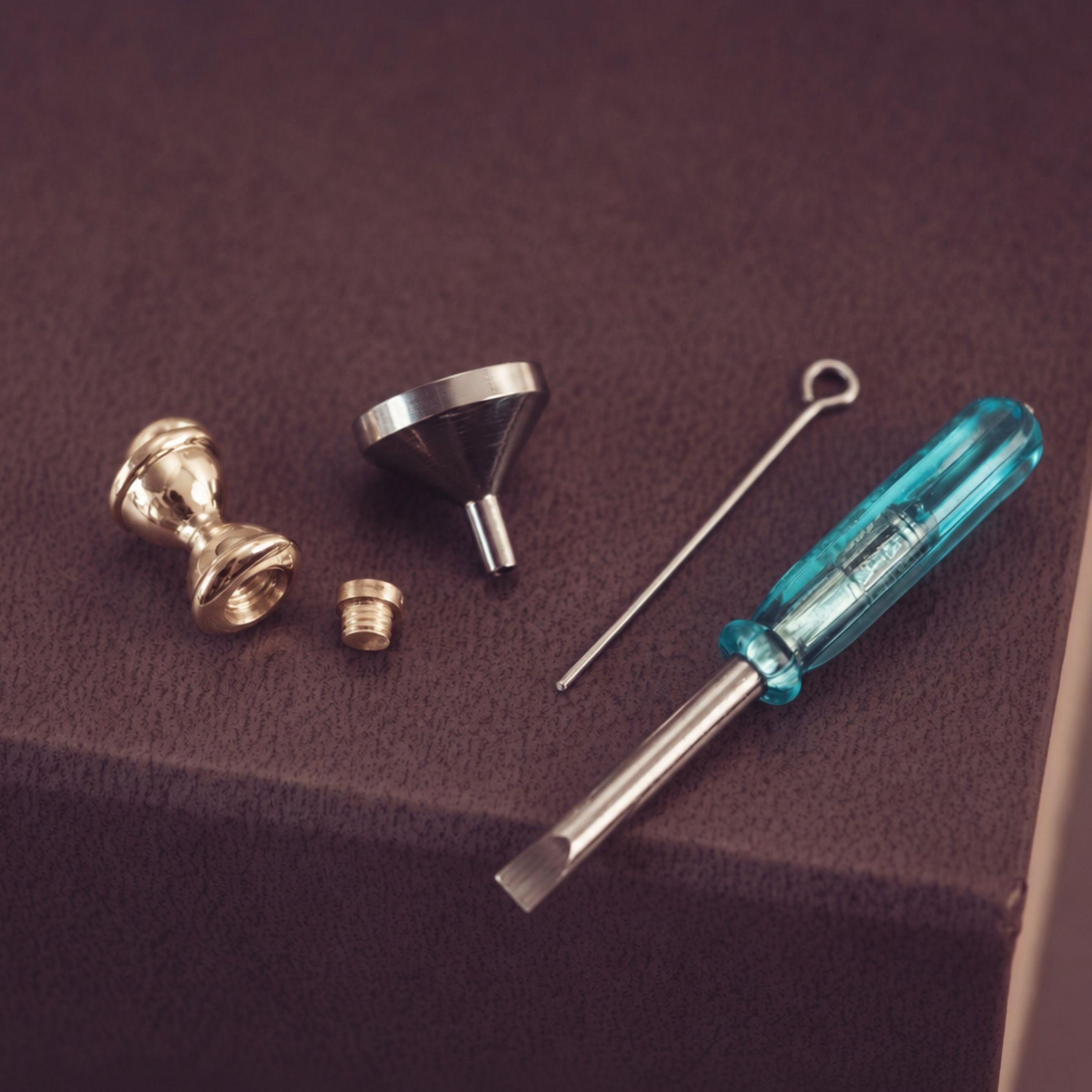 Jewellery tools - a metal funnel, a thin metal tool, and a blue-handled screwdriver on a dark brown textured surface.