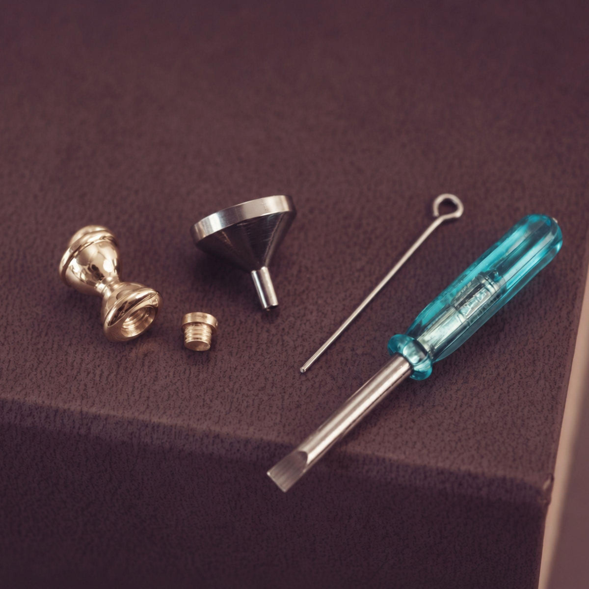 Jewellery tools - a metal funnel, a thin metal tool, and a blue-handled screwdriver on a dark brown textured surface.