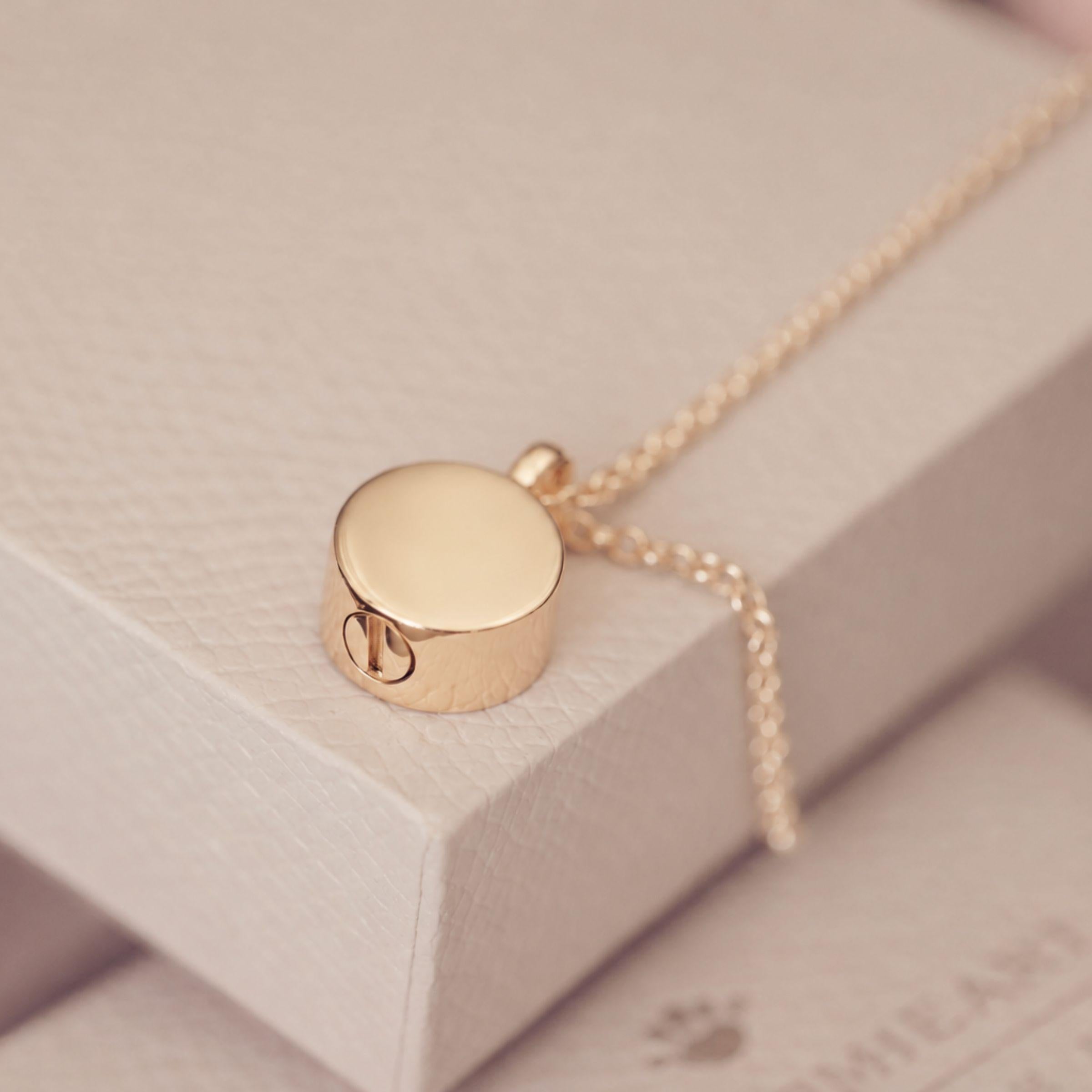 A gold-toned cremation round urn pendant necklace resting on a textured cream-colored jewelry box.