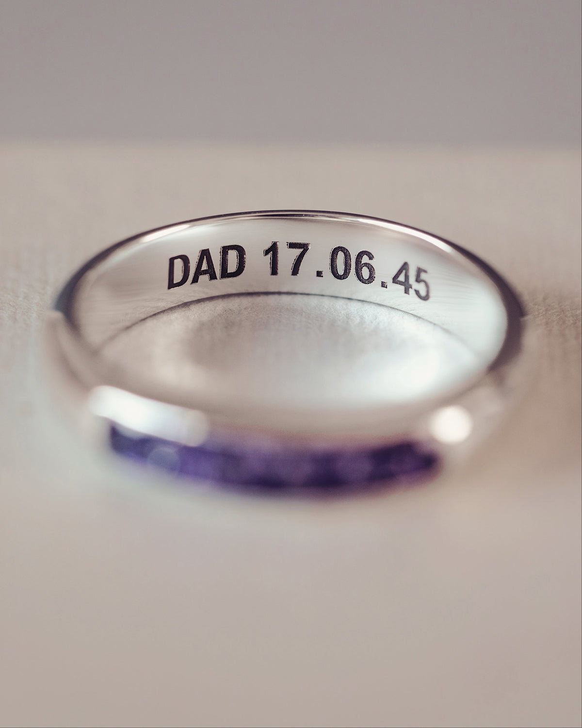 A silver band ring engraved with 'DAD 17.06.45' and set with purple crystals, resting on a textured beige surface.
