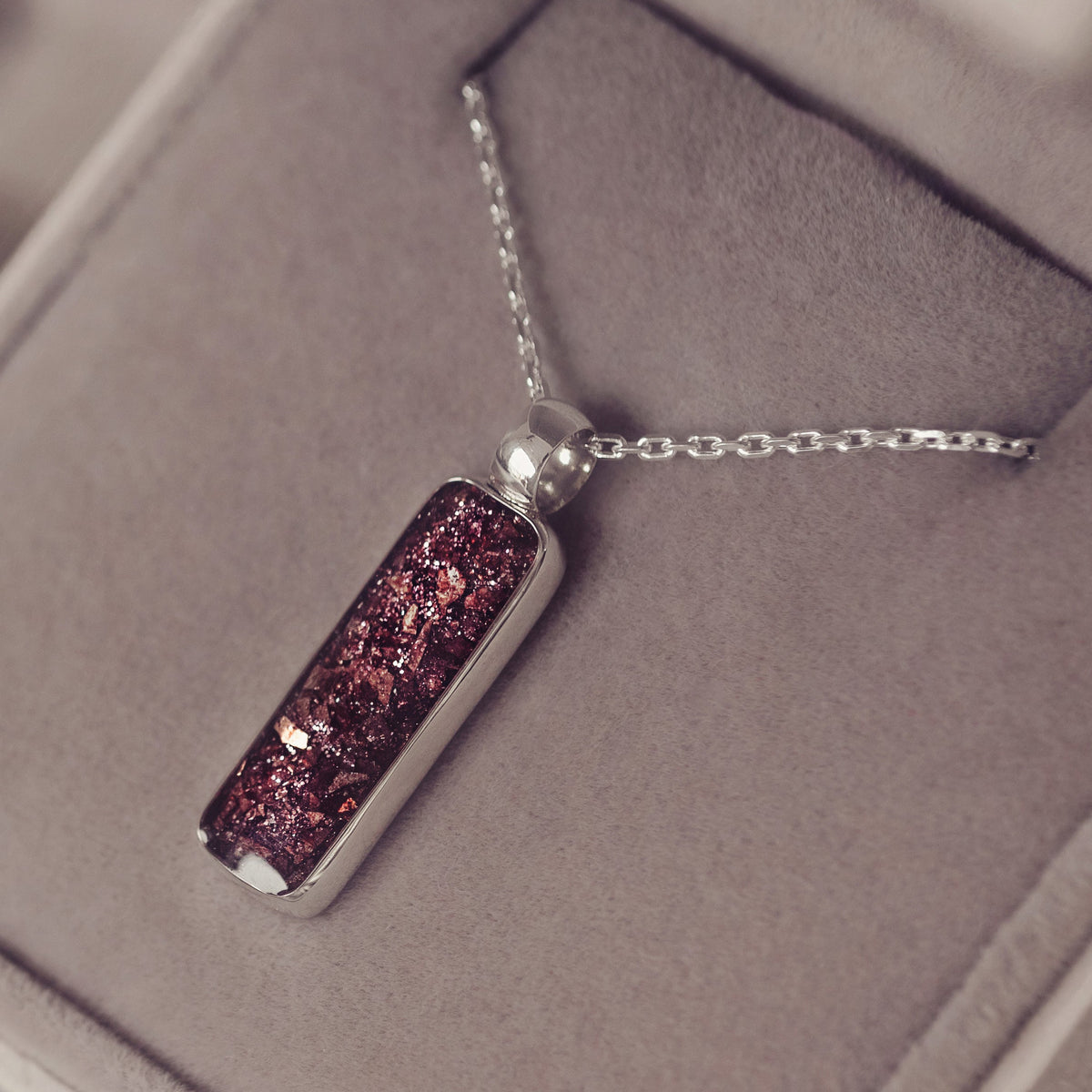 A silver-toned rectangular pendant necklace with a sparkling dark red glitter insert, displayed inside a grey jewellery box on a soft neutral fabric surface.