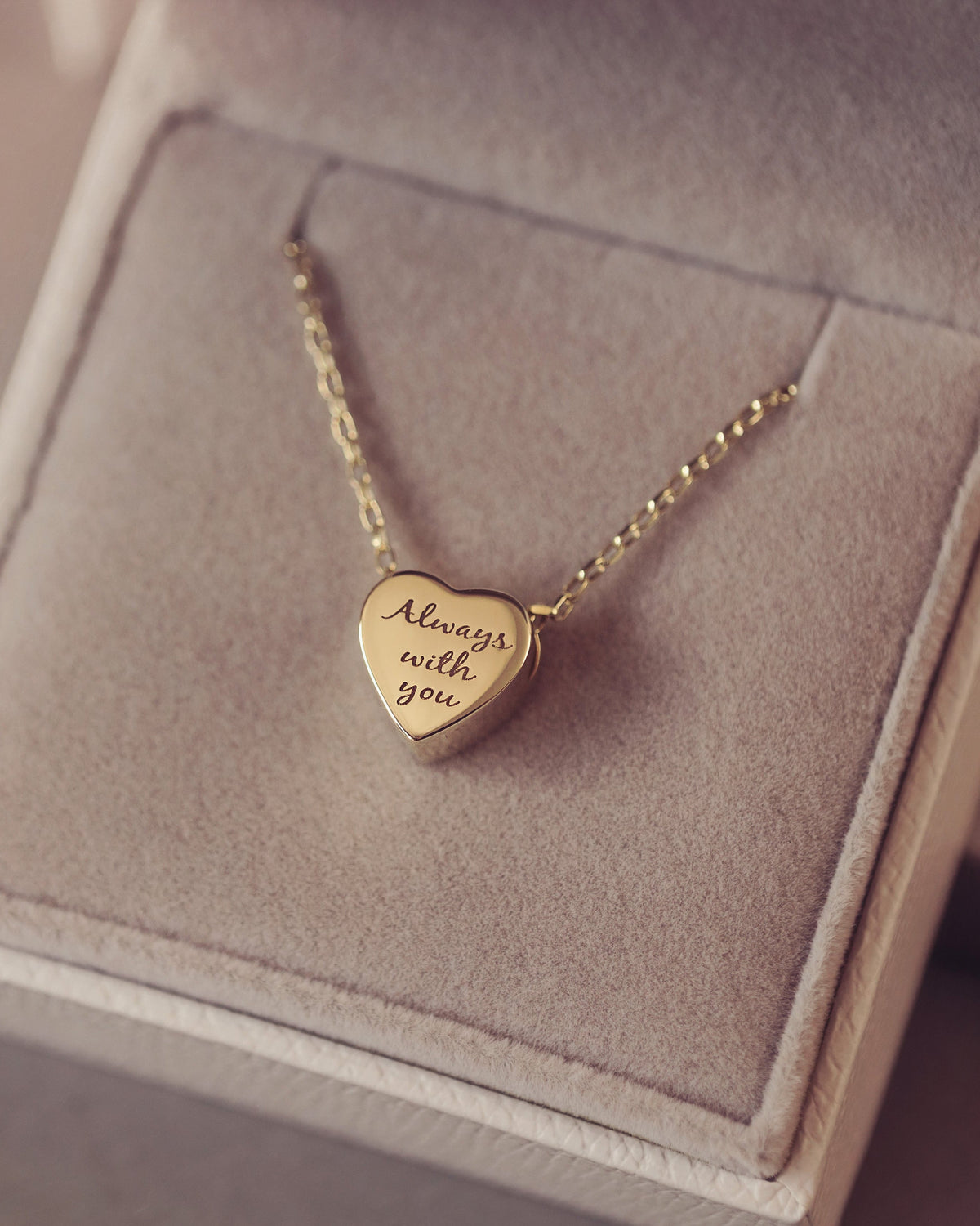 A gold heart pendant necklace engraved with 'Always with you' resting inside a light gray velvet jewelry box.