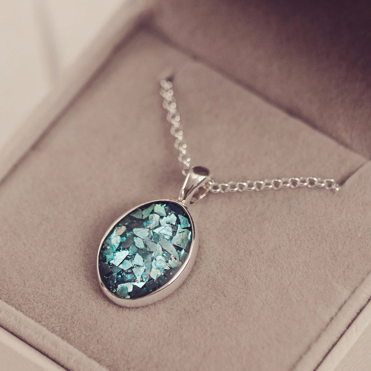 An oval pendant necklace with a sparkling light blue stone in a silver bezel, displayed inside an open beige jewellery box on a soft beige surface.