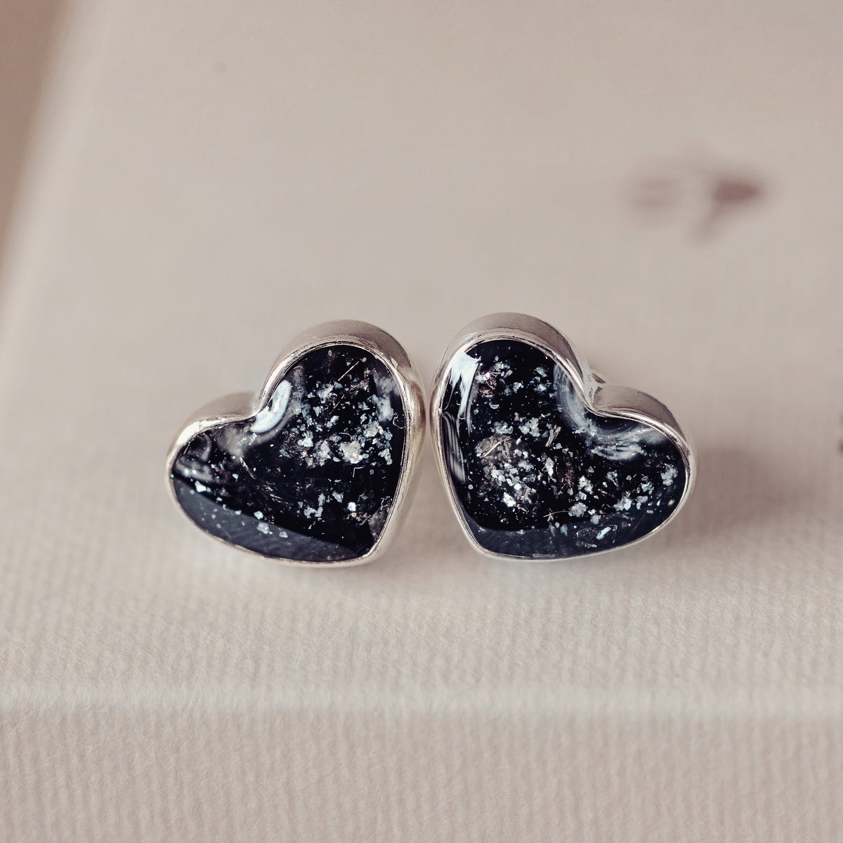 A pair of heart-shaped stud earrings with a dark navy blue glittery inlay and silver borders, resting on a cream-colored jewellery box.