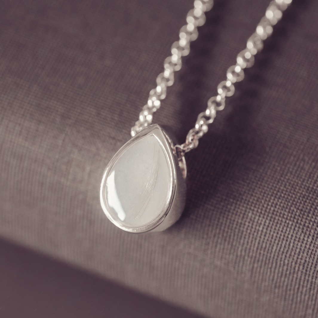 A silver teardrop pendant necklace with a smooth, polished white stone, resting on a textured gray fabric surface.