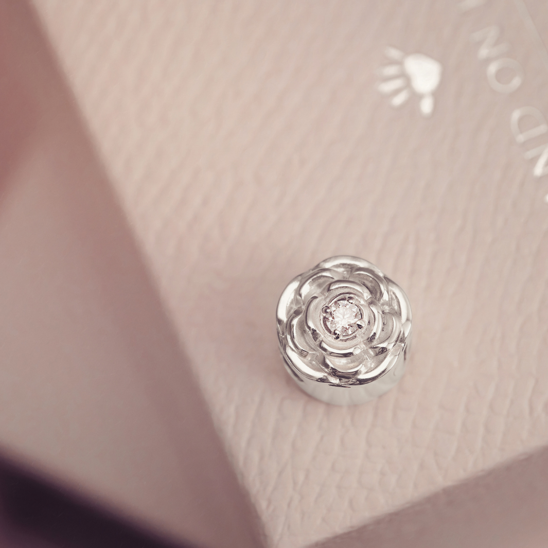A silver-toned rose-shaped charm with a clear central gemstone, resting on a textured light pink surface.
