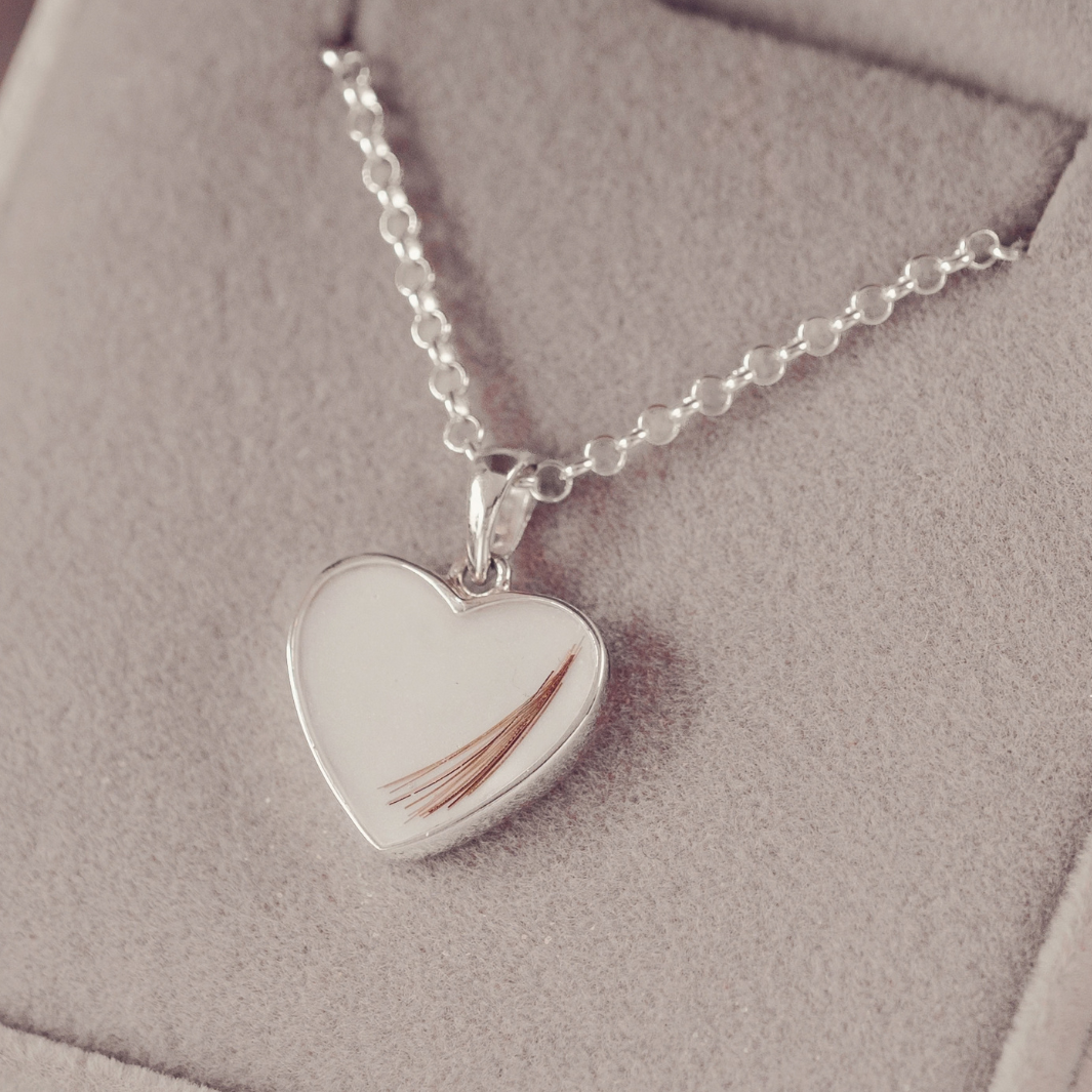 A silver heart-shaped pendant necklace containing a single strand of brown hair, displayed on a soft beige jewelry box.