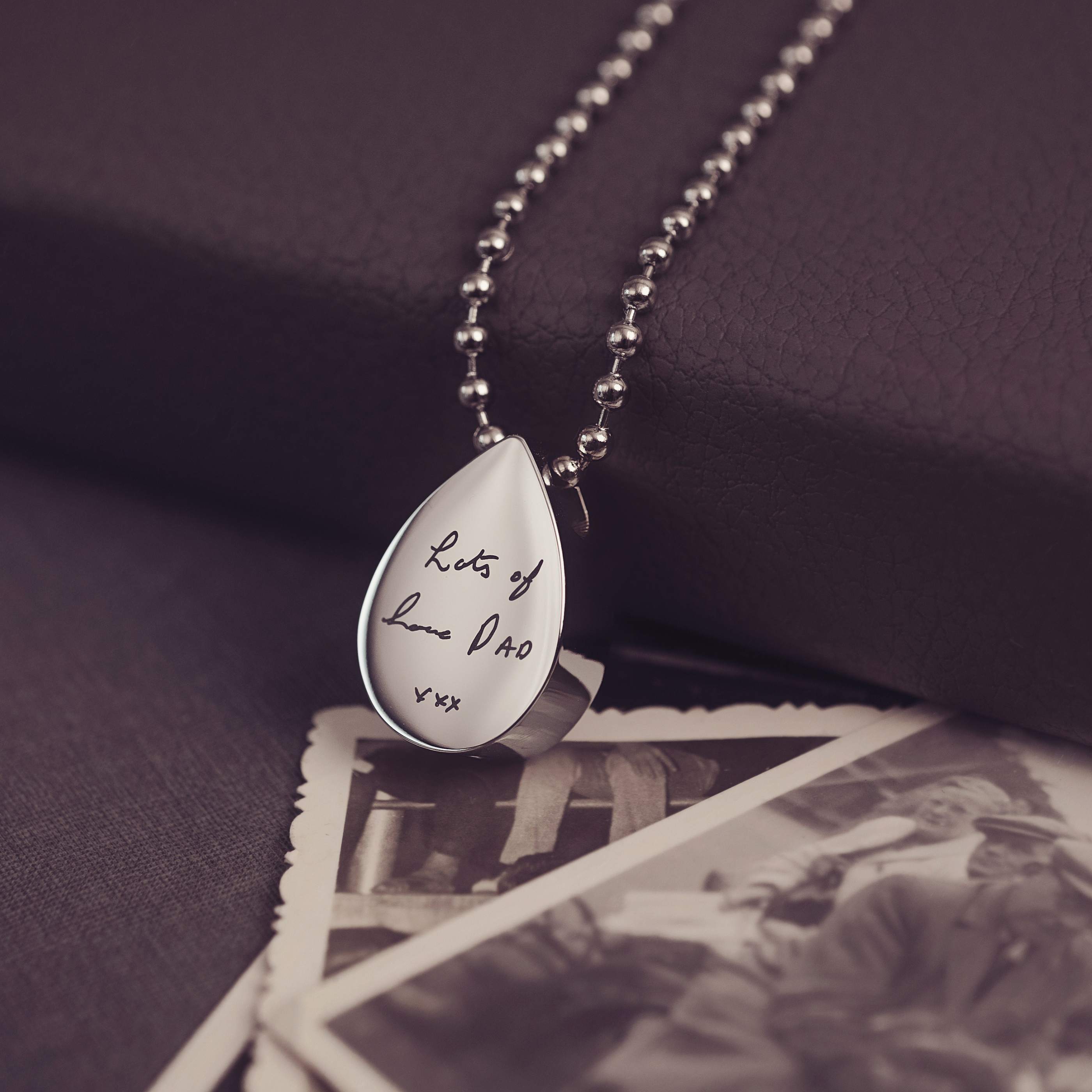 Teardrop-shaped silver pendant with engraved text on a dark background with vintage photos.