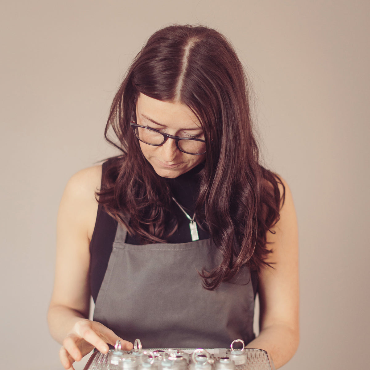 A woman with long dark hair and glasses, wearing a gray apron over a black top, holds a metal tray containing several small silver rings against a plain beige background.