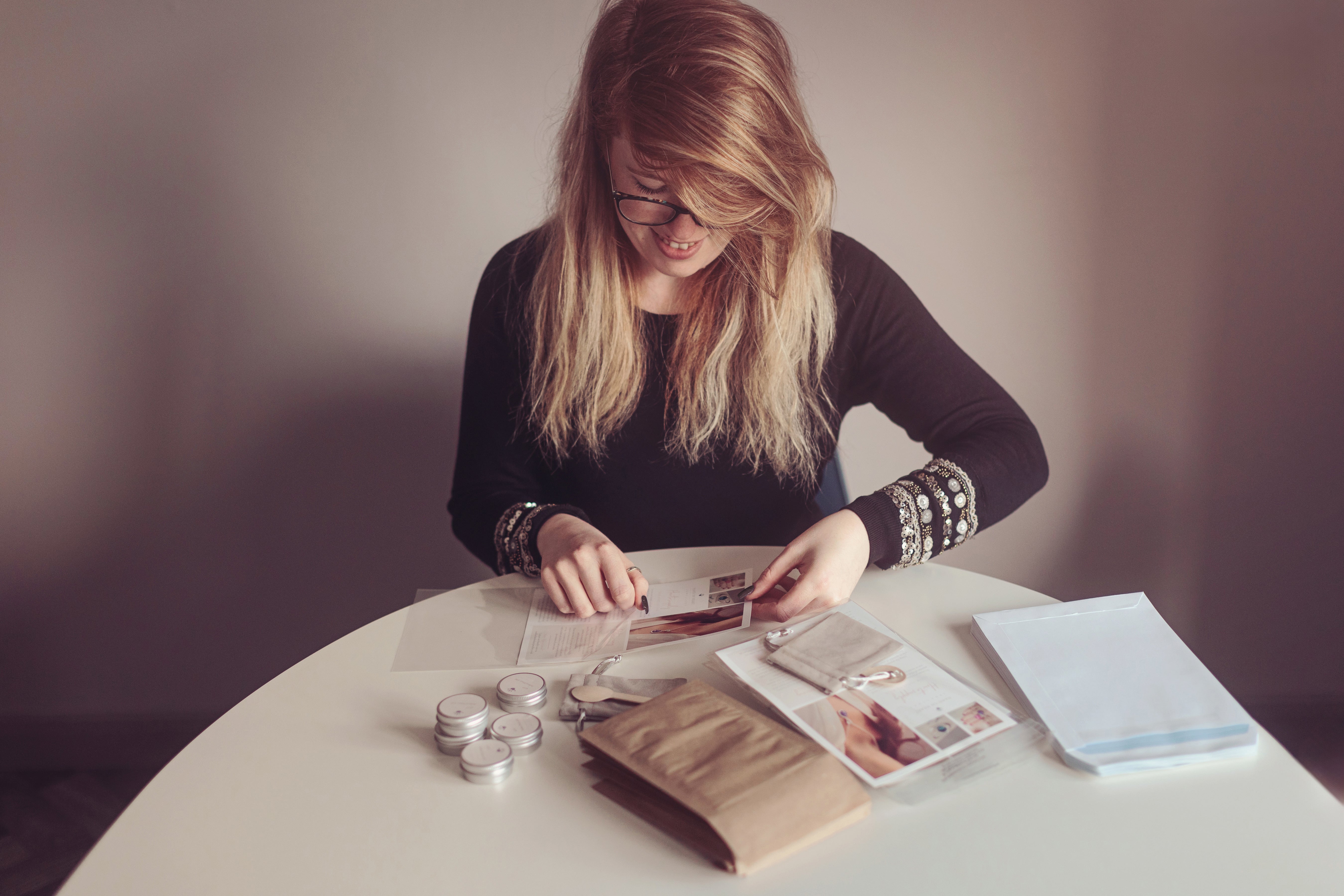A woman with long blonde hair and glasses sits at a white round table, looking down at a small package she is holding. On the table are several small round silver jars, a brown paper bag, a booklet, and a stack of white envelopes or mailers against a plain, warm-toned wall.