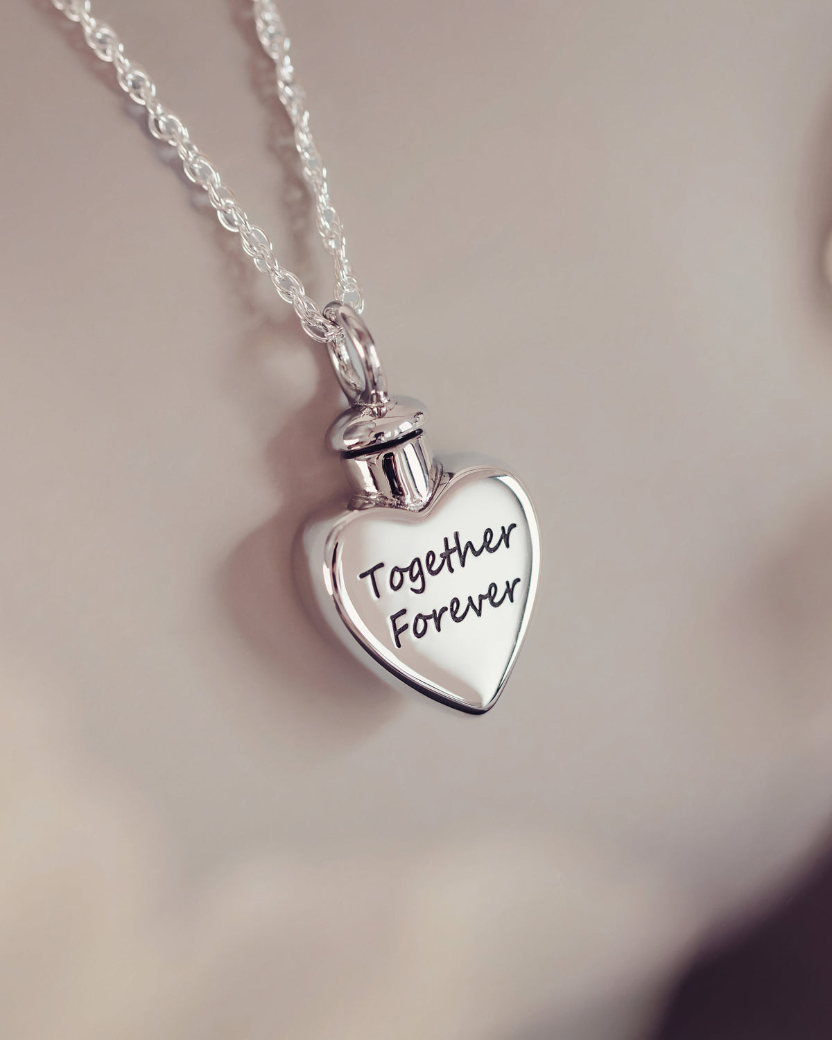 A silver heart-shaped cremation urn pendant engraved with 'Together Forever', hanging from a delicate silver chain against a soft, warm beige background.