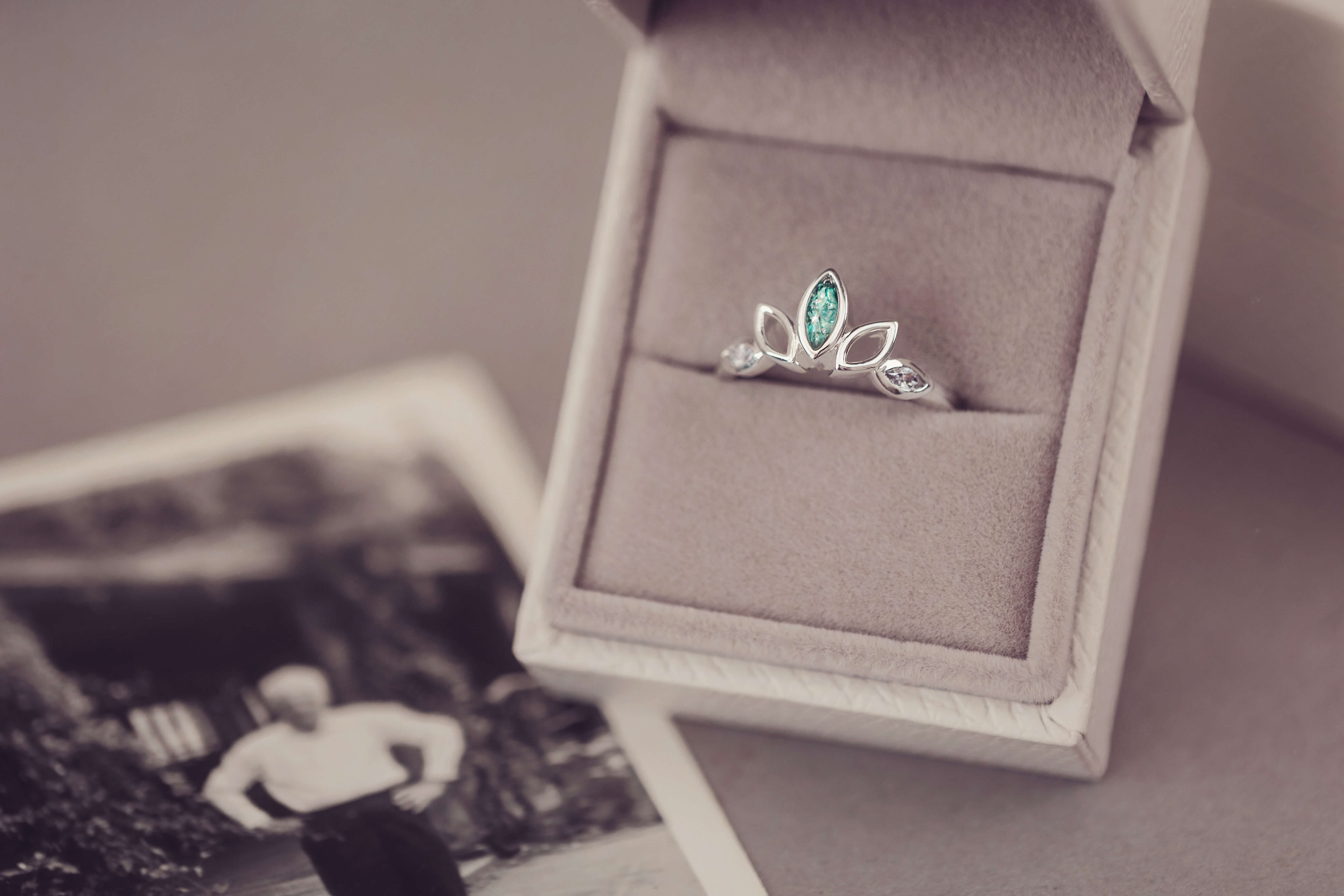 Sterling Silver Ring, created with your loved ones ashes, with Cubic Zircona gemstones. Green floating crystals with in piece in a grey jewellery box placed on top of a blurred photo 