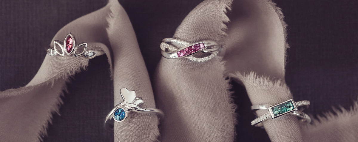 Four silver rings with colorful gemstones are displayed on a draped beige fabric against a dark background.