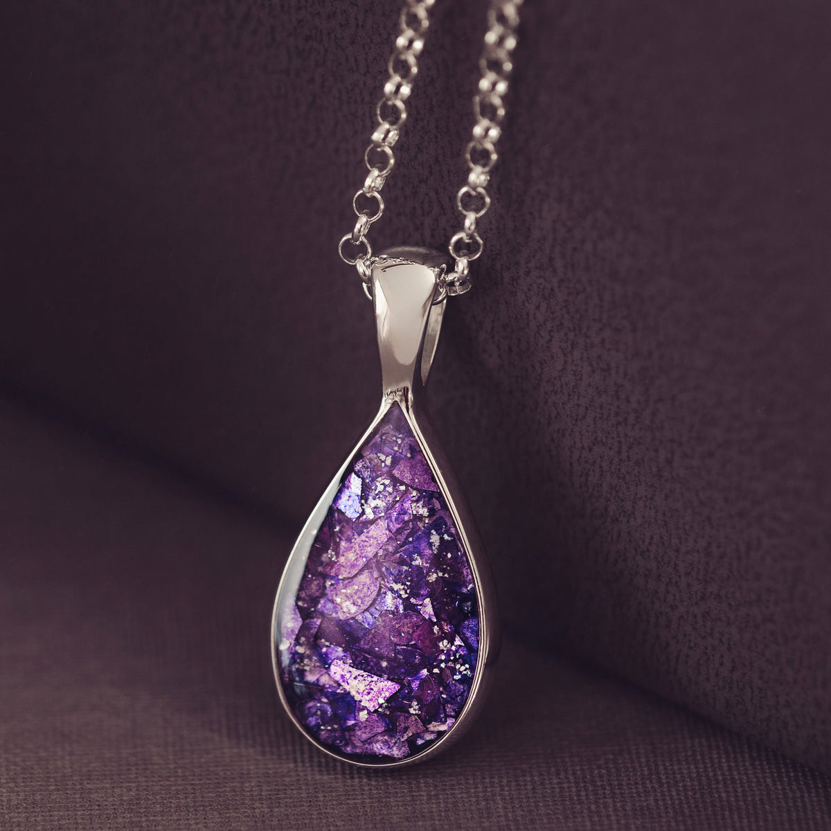 A teardrop-shaped pendant necklace with a sparkling purple and silver glitter design, set in a silver-toned bezel and hanging from a silver chain, displayed on a dark brown textured surface.