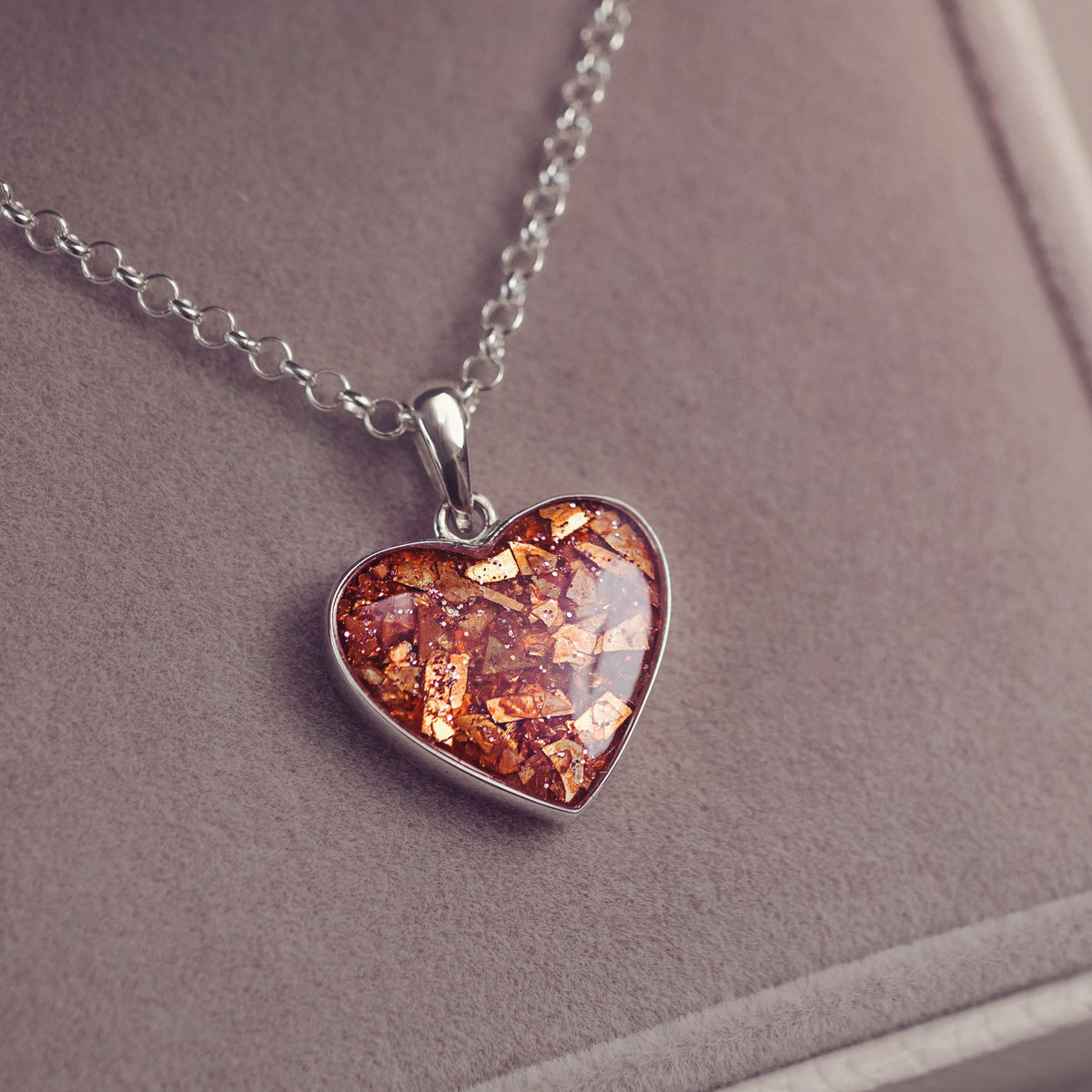 A silver heart pendant necklace with a textured amber-orange inlay, displayed inside a grey velvet jewellery box.
